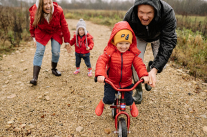 Kuvassa pieni lapsi opettelee ajamaan polkupyörällä, mukana toinen lapsi ja kaksi aikuista, jotka kannustavat häntä