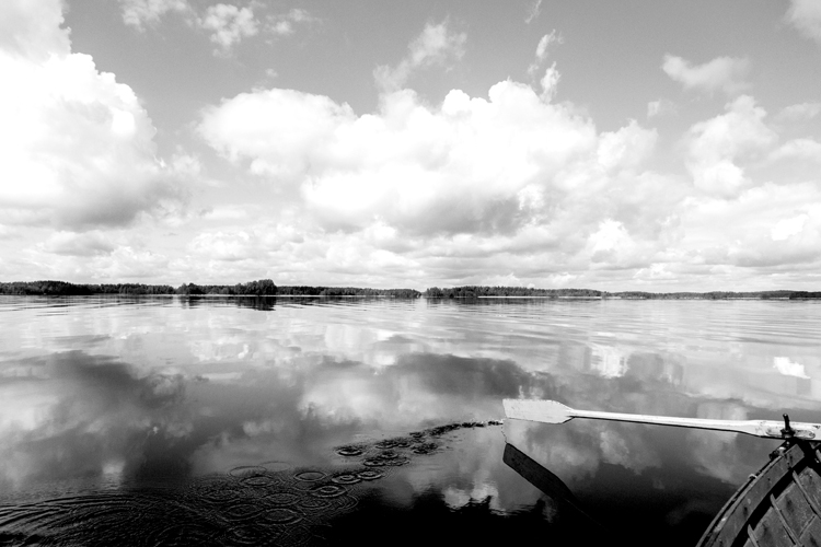 Mustavalkokuvassa peilityyni järven pinta veneestä kuvattuna, etualalla airo.