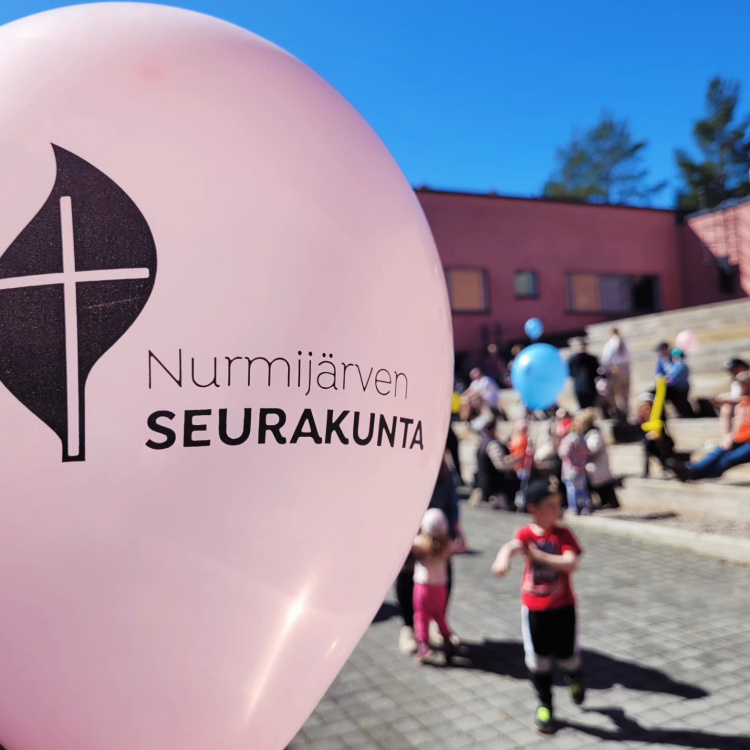 Klaukkalan Vappu Kuvassa edessä seurakunnan logolla varustettu ilmapallo. lapsia taka-alalla